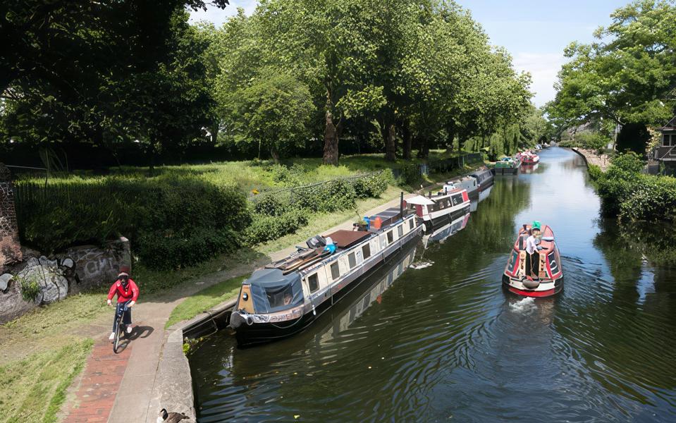 A high angle photograph of narrowboats moored on the canal. A cyclist passes on the towpath