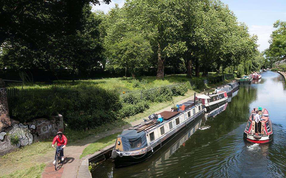 A high angle photograph of narrowboats moored on the canal. A cyclist passes on the towpath