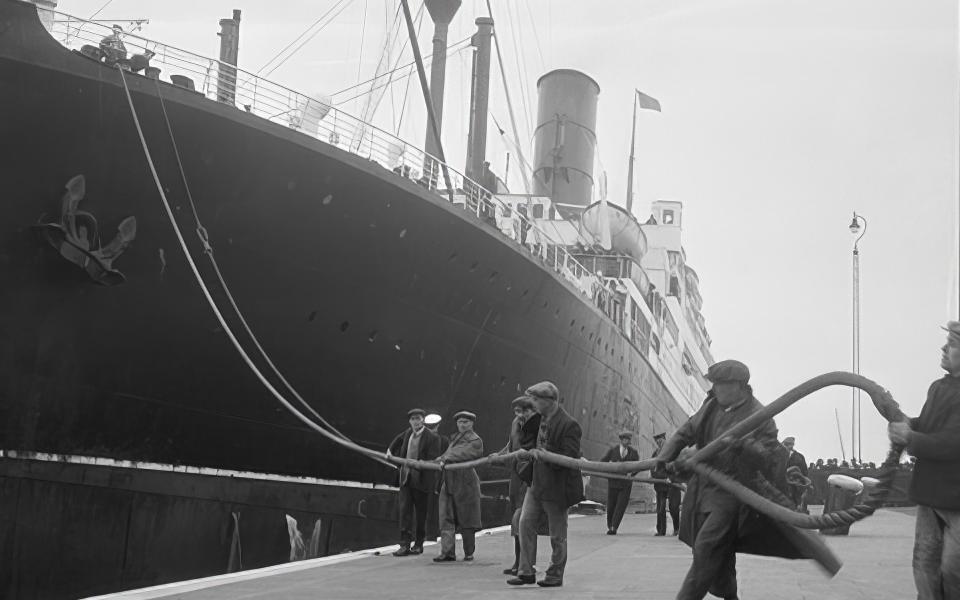Monochrome photograph of men on a dockside holding one of the mooring ropes of a huge ocean liner