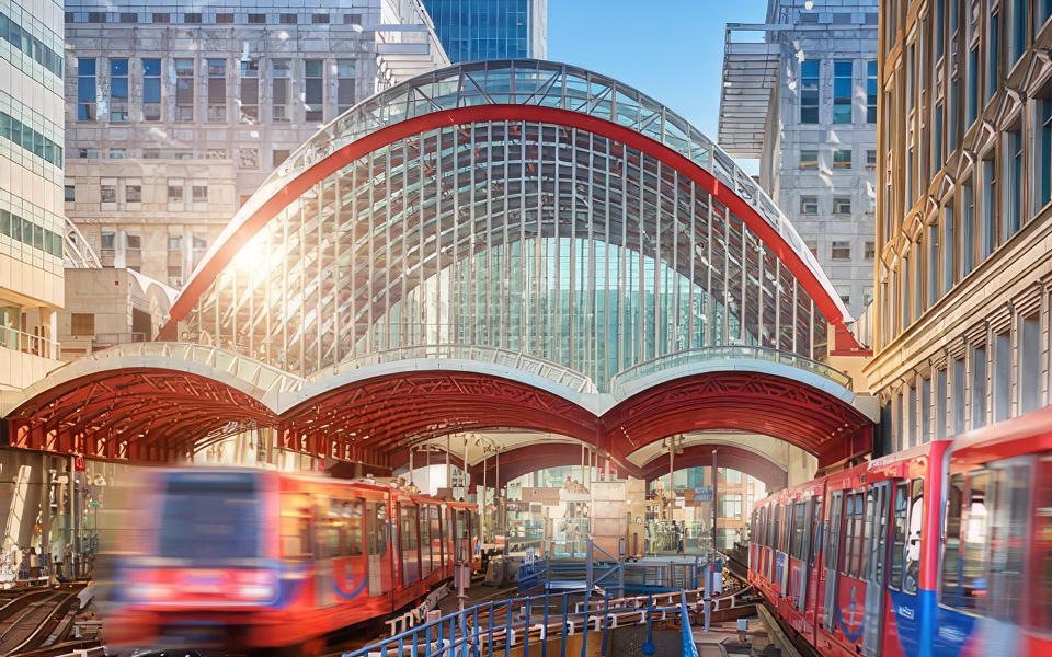 An action photo of Docklands light Railway trains moving towards the camera under a set of arching glass structure